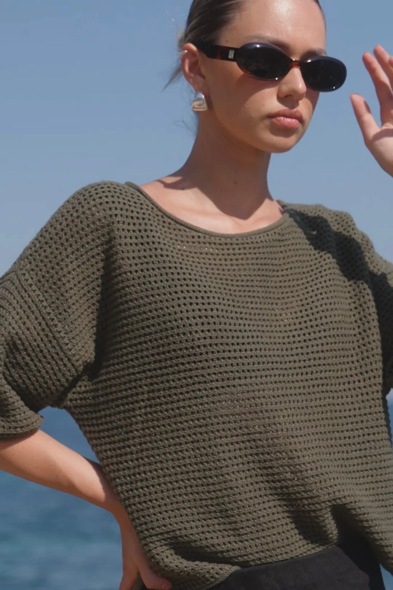 Woman wearing Re:Union The Label Sunrise Khaki SS Crochet Knit Top in crochet knit with short sleeves, relaxed fit, and round neckline, styled with denim jeans.